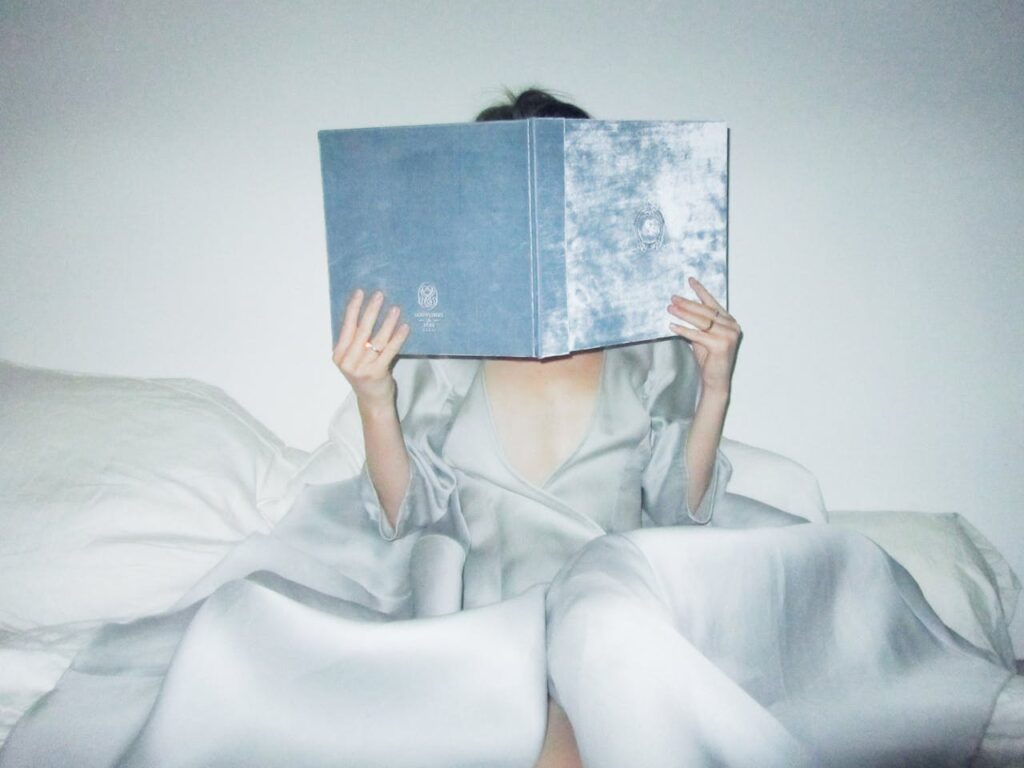 Photo of a woman in bed. She is holding up a large, light blue book. Her face is hidden behind the large book.