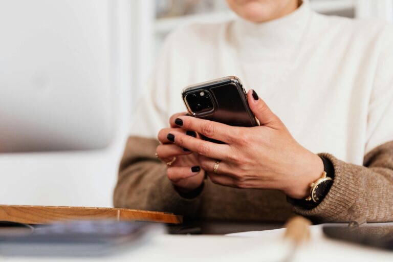 Photo of a woman, but only of her torso and hands. She is holding a cell phone and has black fingernail polish. She is wearing a white and brown sweater.