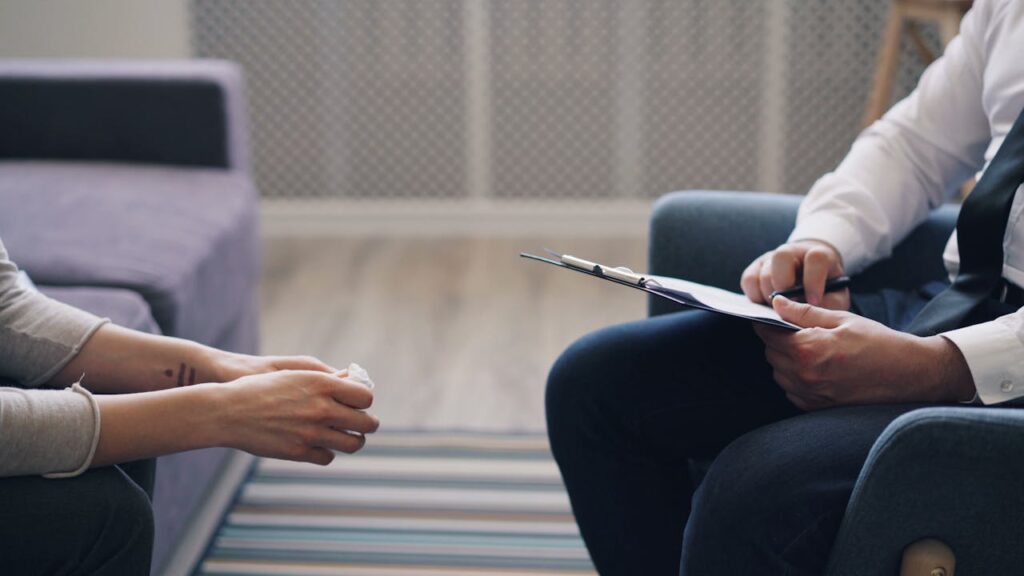 Photo of two people sitting across from each other. One person has a clipboard, and the other has a tissue in their hand.